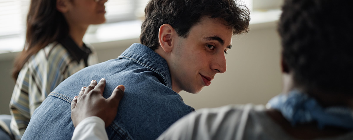 Ein junger Mann sitzt in einer Gruppe und wird von einer Person neben ihm beruhigend an der Schulter berührt.